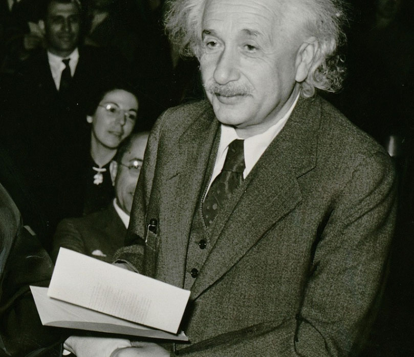 Albert Einstein holding an envelope. An audience can be seen in the background.