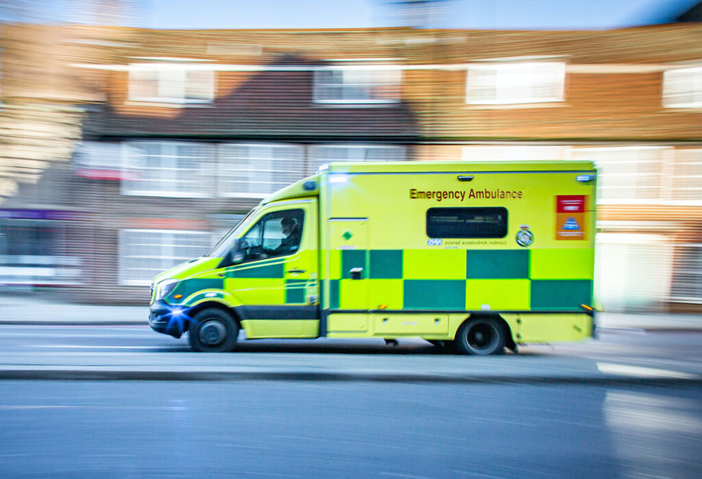 Side view of a bright yellow ambulance with green flashes driving along a road. In the background is a row of houses. The houses are blurred because the ambulance is moving so fast.