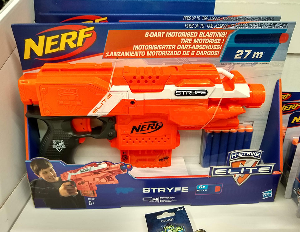 A Nerf Gun Stryfe in packaging on a shop shelf. There is a picture of a boy holding a Nerf Gun on the packaging.