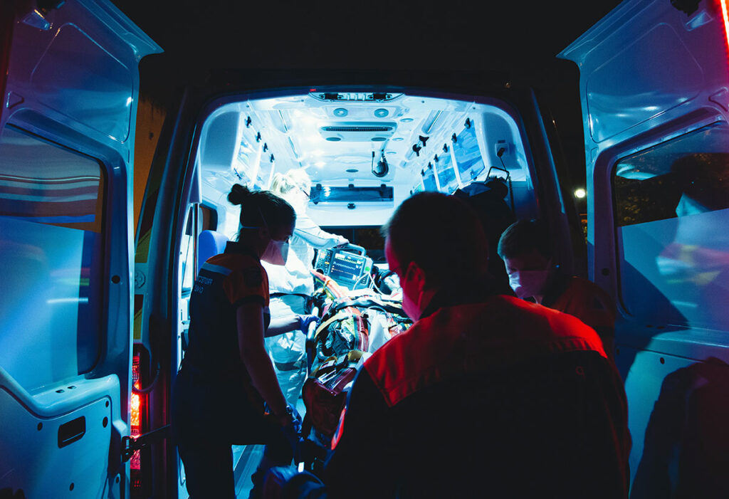 Paramedics work in an ambulance. It is night time and the interior of the ambulance is very bright. Three paramedics stand in the foreground in darkness.
